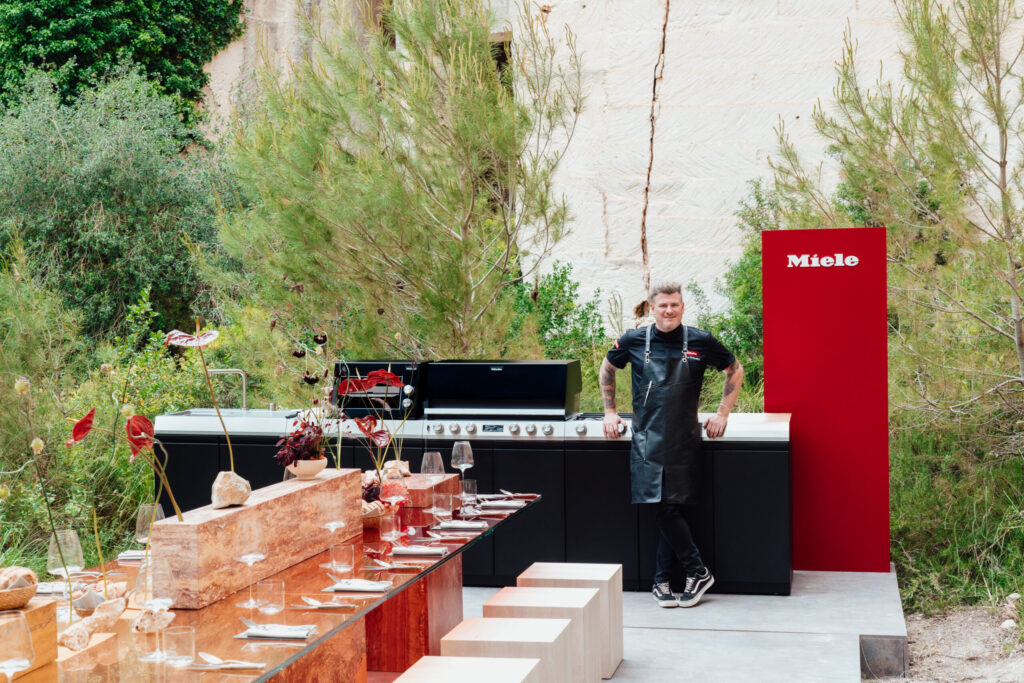Koch neben Miele Outdoor-Küche Dreams beim Launch-Event auf Menorca, roter Miele-Aufsteller im Hintergrund