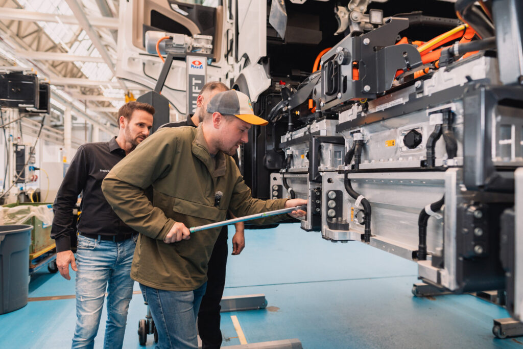 Drei Techniker inspizieren Hochvolt-Batteriemodule an einem aufgebockten Elektro-Lkw in einer Werkstatt