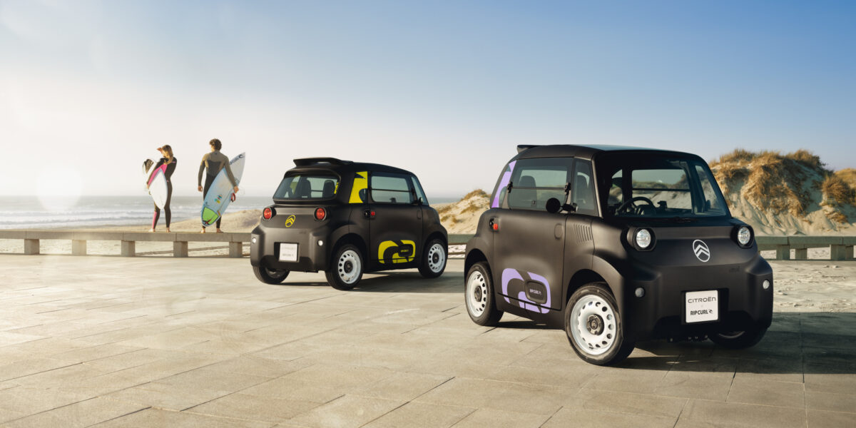 Zwei Citroën Ami Rip Curl Sondermodelle auf einer Strandpromenade mit Surfern im Hintergrund