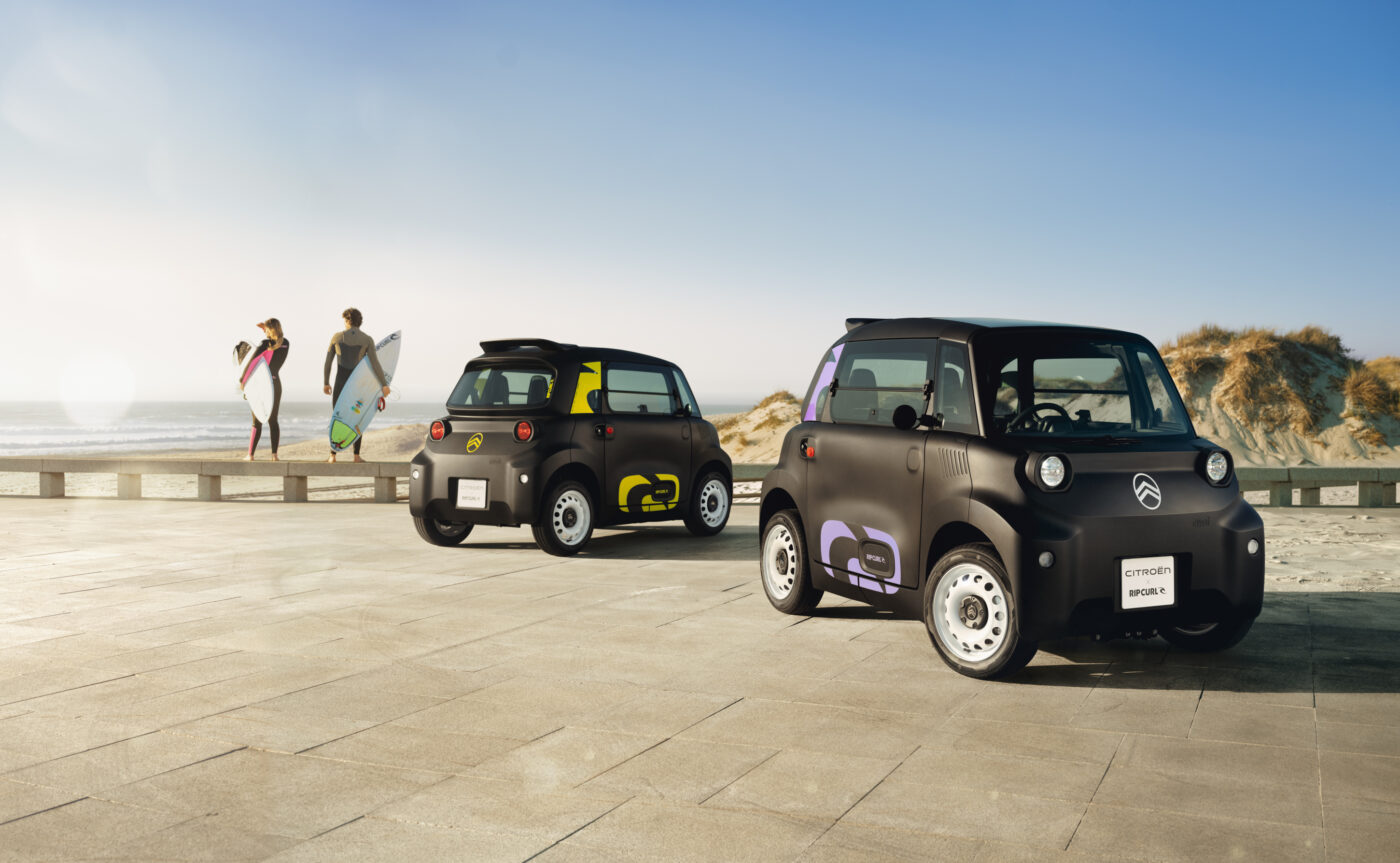 Zwei Citroën Ami Rip Curl Sondermodelle auf einer Strandpromenade mit Surfern im Hintergrund