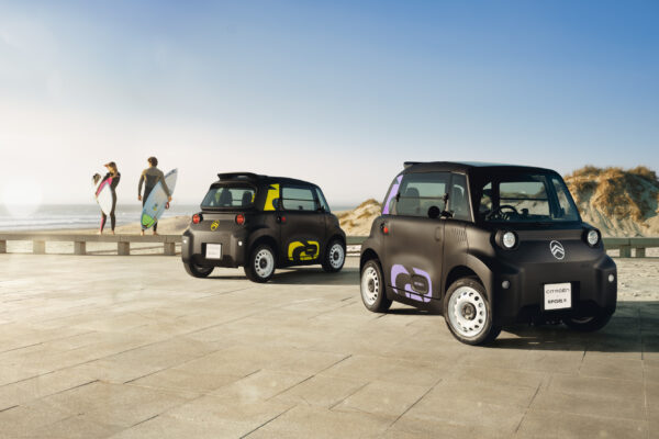 Zwei Citroën Ami Rip Curl Sondermodelle auf einer Strandpromenade mit Surfern im Hintergrund