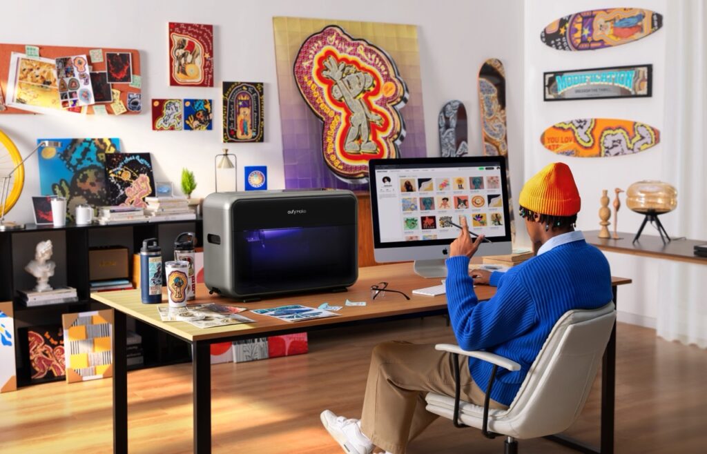 eufyMake E1 UV-Drucker auf Schreibtisch, daneben Person am Apple iMac in buntem Kreativstudio