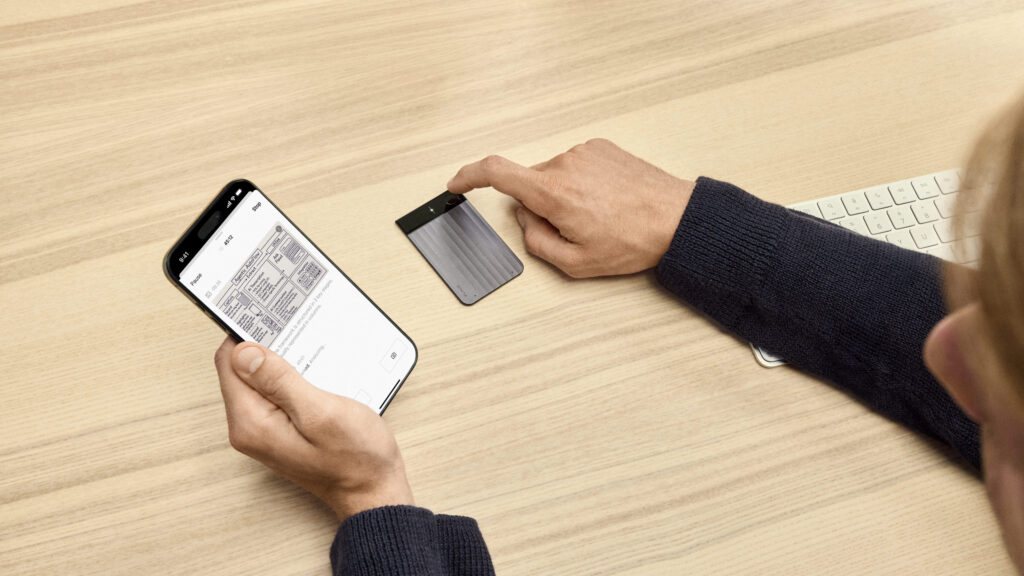 Hand tippt auf Plaud Note KI-Diktiergerät auf Holztisch, daneben iPhone mit geöffneter App