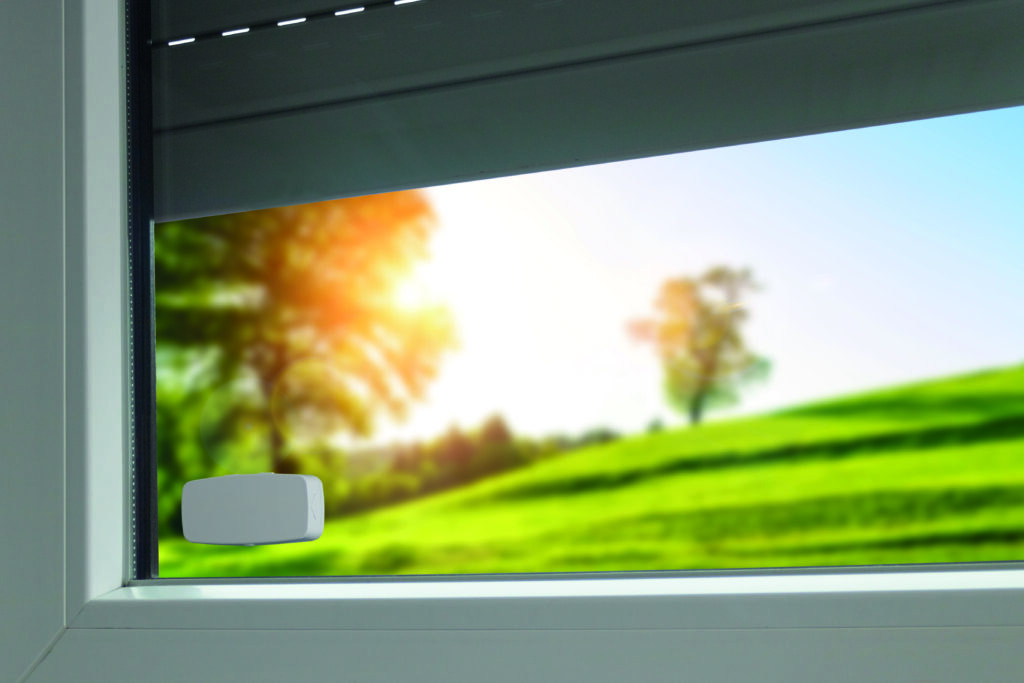 Rademacher DuoFern-Sensor am weißen Fensterrahmen, darüber halb geschlossener Rollladen bei Sonnenschein