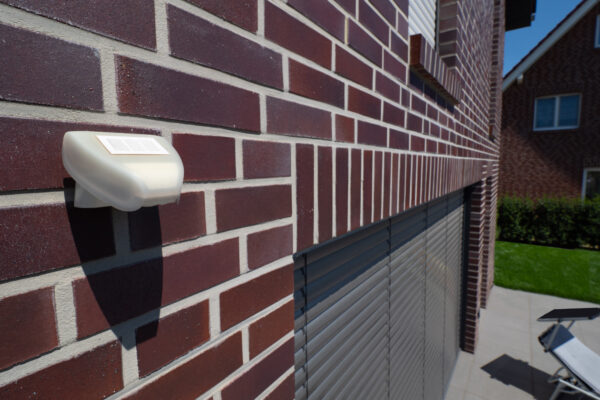 Rademacher Sonnensensor an Klinkerfassade montiert, dahinter Rollladen und Terrasse