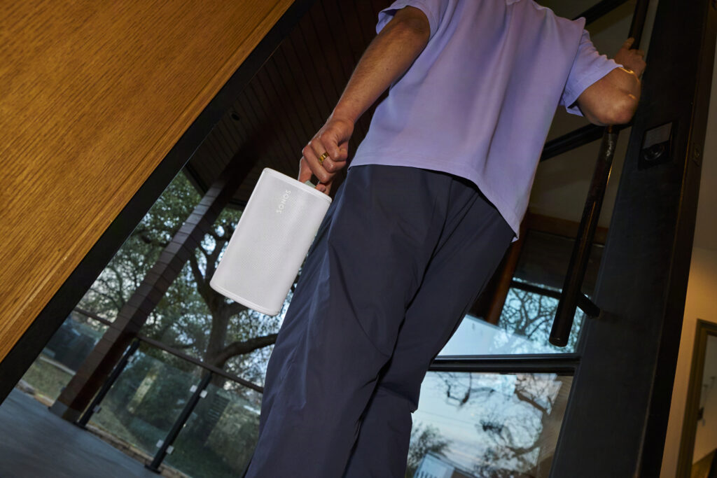 Person trägt weißen Sonos-Lautsprecher in der Hand an einer Glastür im Innenbereich