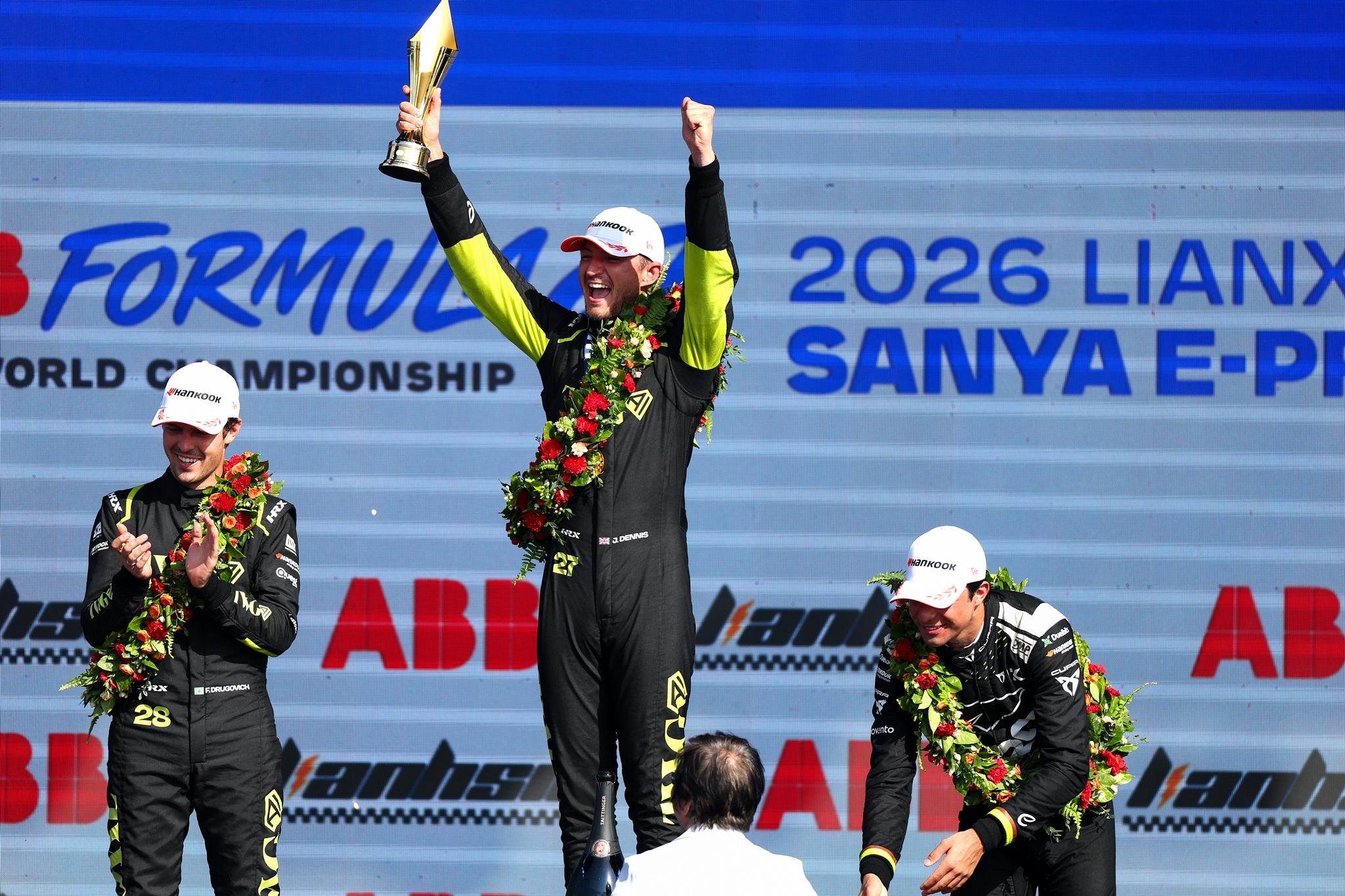 Jake Dennis hebt Siegerpokal beim ABB Formula E Sanya E-Prix, flankiert von Drugovich und einem weiteren Fahrer