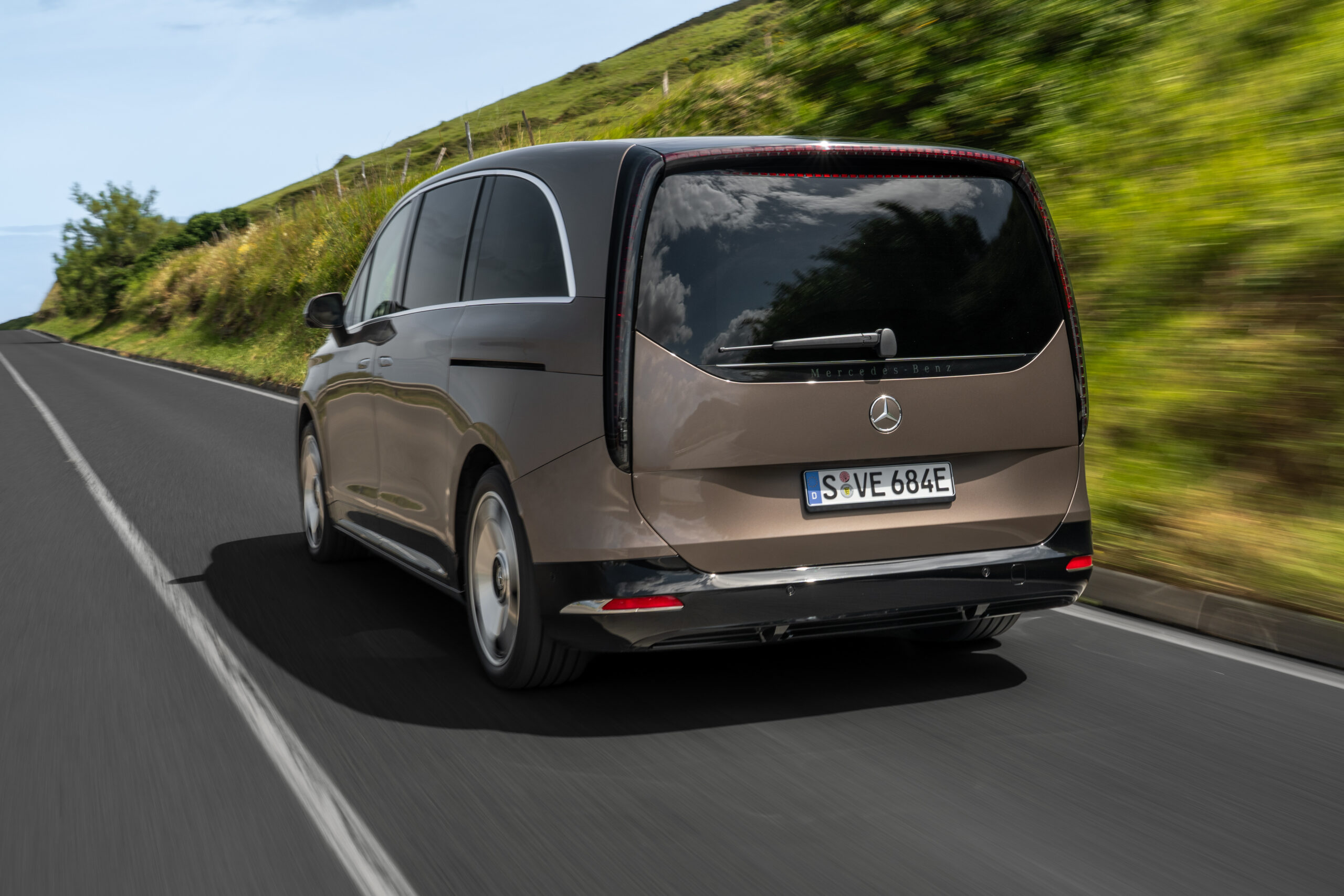 Mercedes-Benz VLE in Bronze-Metallic, Heckansicht auf Landstraße mit grüner Hügellandschaft