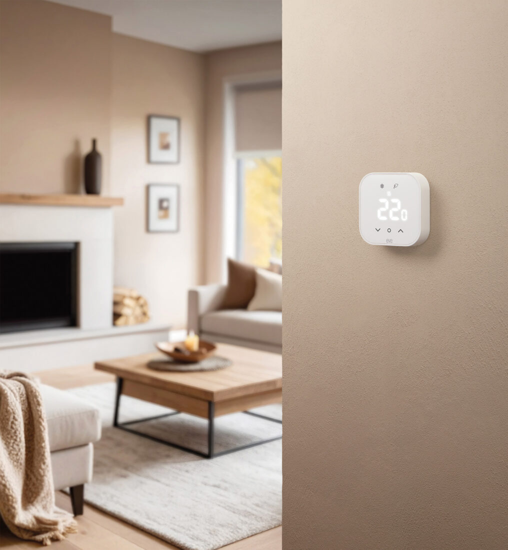 Eve Thermostat an beiger Wand montiert, Display zeigt 22 °C, modernes Wohnzimmer im Hintergrund