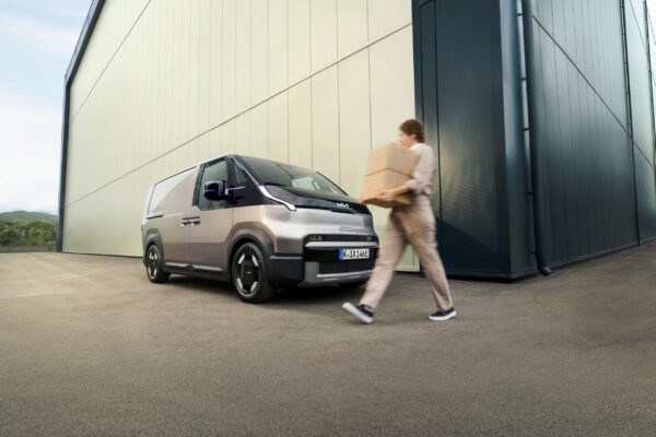 Kia Elektro-Van in Silber vor Lagerhalle, Person trägt Pakete vorbei