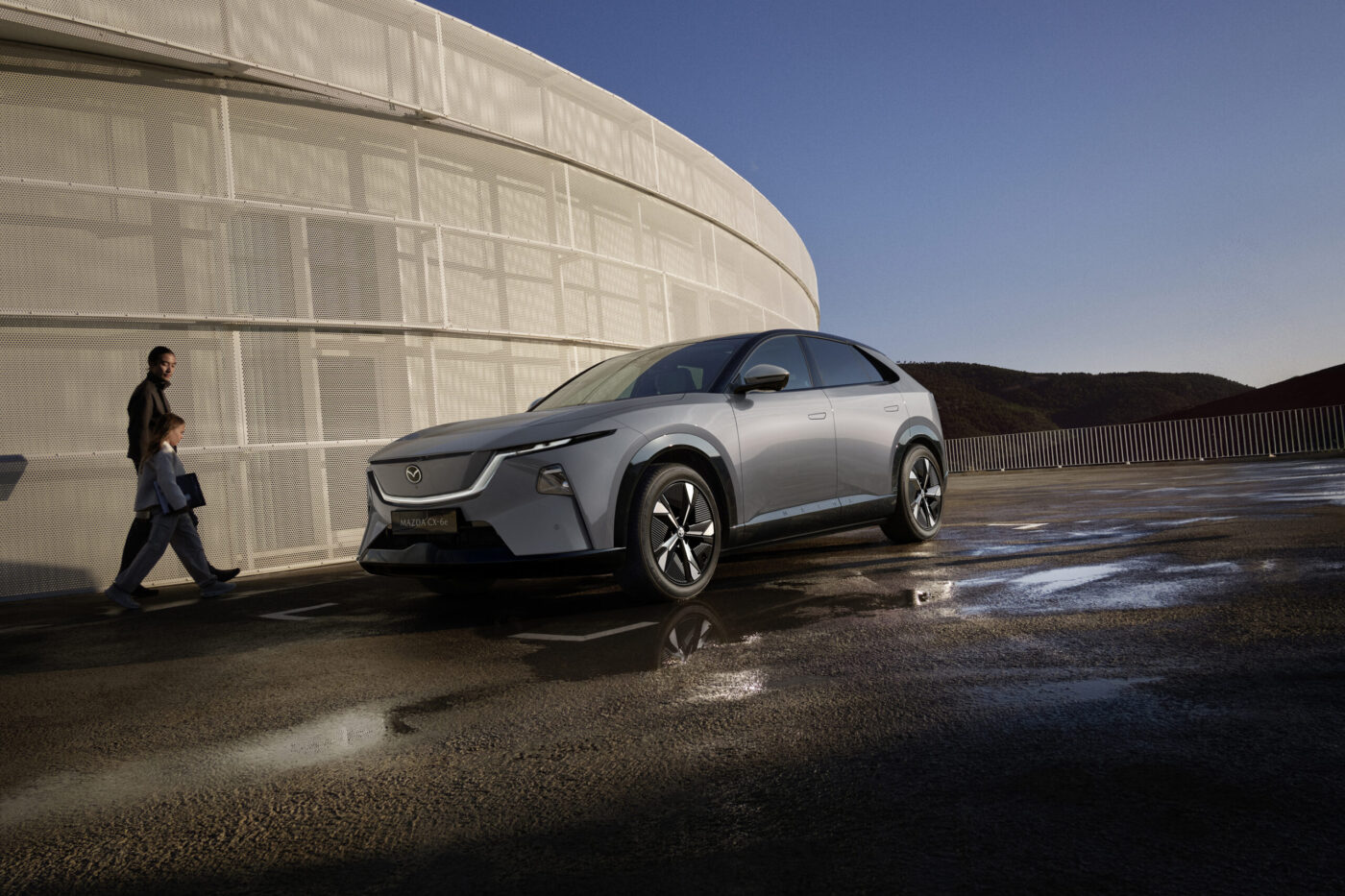 Mazda CX-6e in Silber, Frontansicht schräg links, parkend auf nassem Platz vor Rundbau