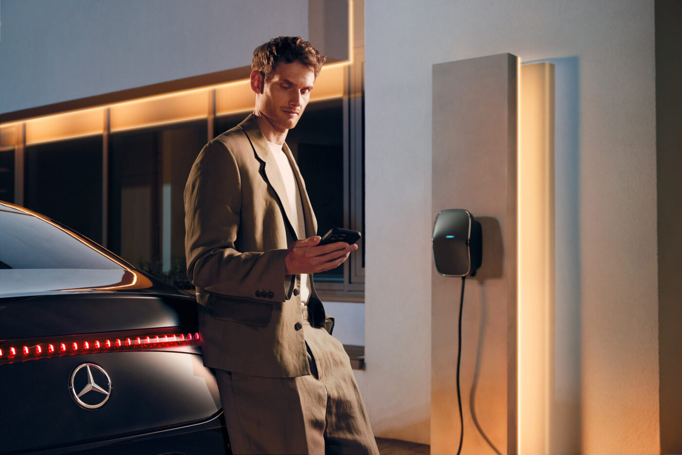 Mann mit Smartphone lehnt an Mercedes-Benz EV neben MB.CHARGE Home Wallbox beim Heimladen
