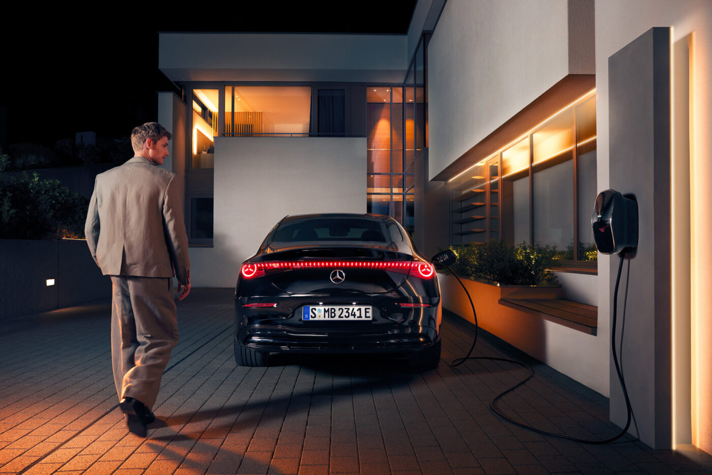 Mercedes-Benz Elektrolimousine wird nachts an Heim-Wallbox geladen, Mann im Anzug nähert sich