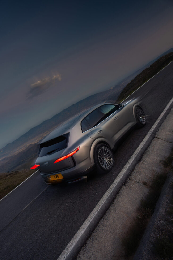 Bentley Torcal in Silbergrau auf Bergstraße bei Dämmerung, Heckansicht mit leuchtenden Rücklichtern