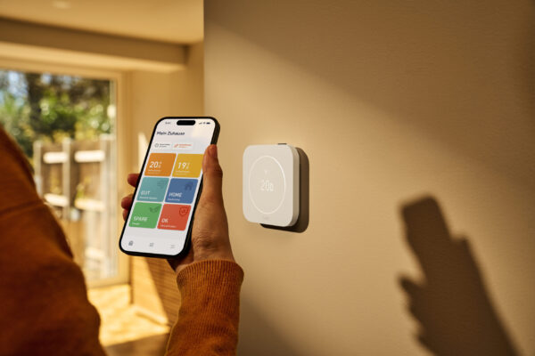 Hand hält Smartphone mit tado°-App neben an der Wand montiertem tado° Thermostat X (2. Gen.)
