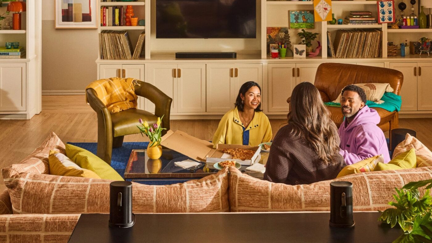 Drei Personen auf Sofa mit Pizza, flankiert von zwei tragbaren Sonos Surround-Speakern, Soundbar unter TV