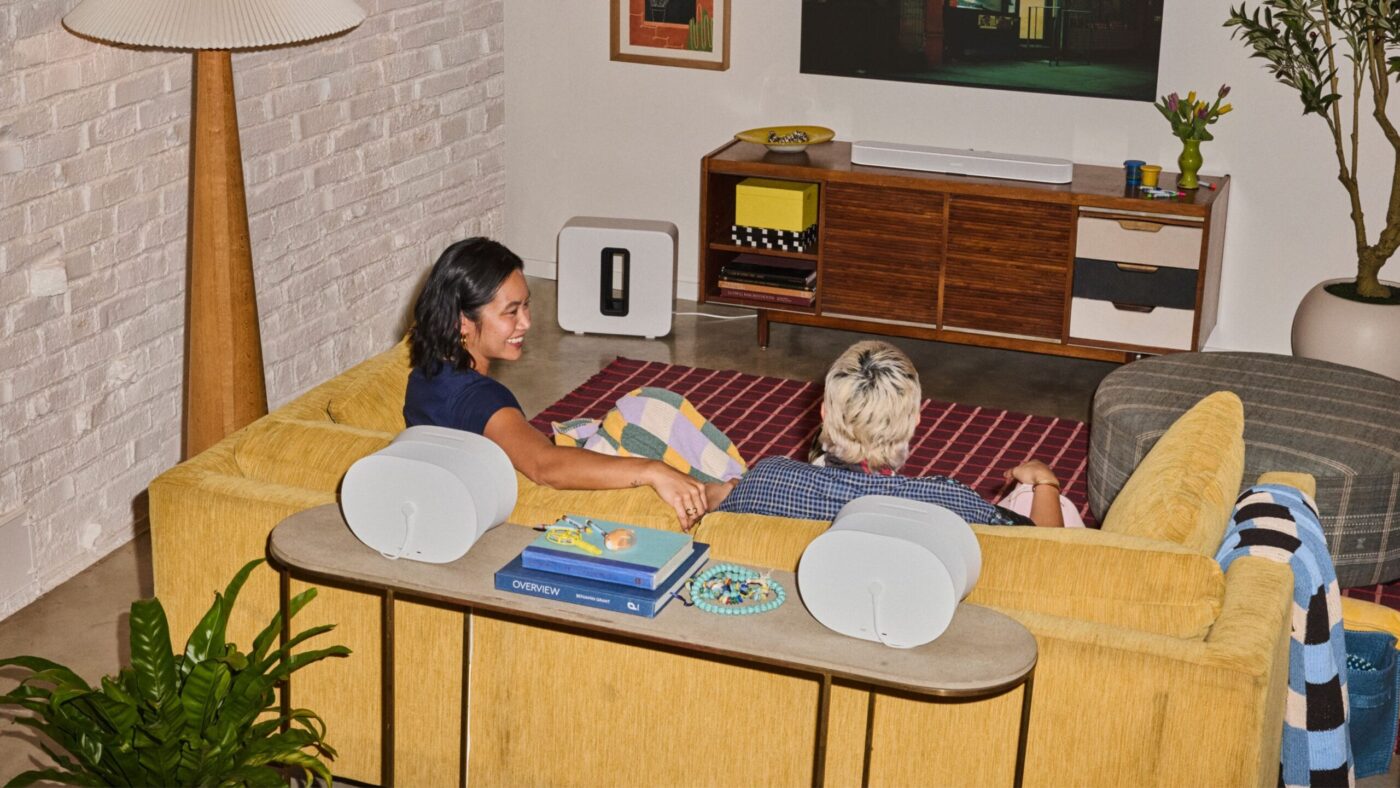 Zwei Personen auf gelbem Sofa mit zwei Sonos Era 300 als Surround-Lautsprechern und Sonos Sub im Wohnzimmer