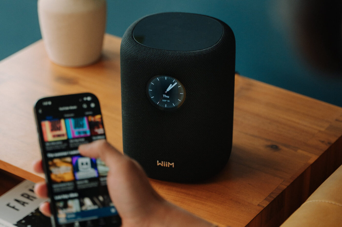 WiiM Smart Speaker mit Display auf Holztisch, gesteuert über die WiiM Home App auf einem Smartphone