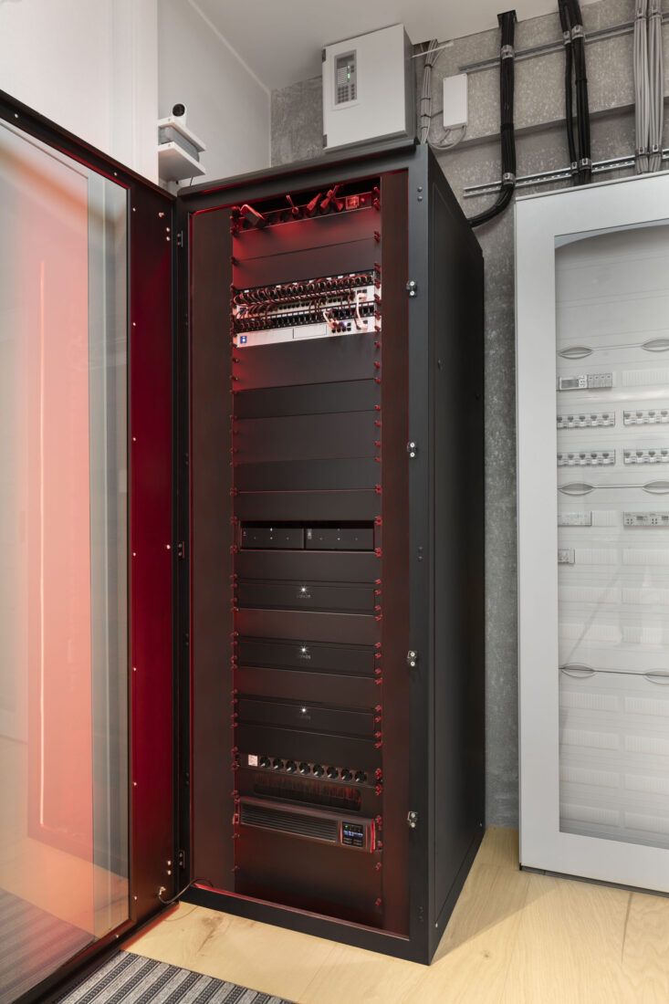 Geöffnetes schwarzes AV-Rack mit rot beleuchteten Einschüben, u. a. Sonos Amp, im Technikraum eines Berliner Penthouses