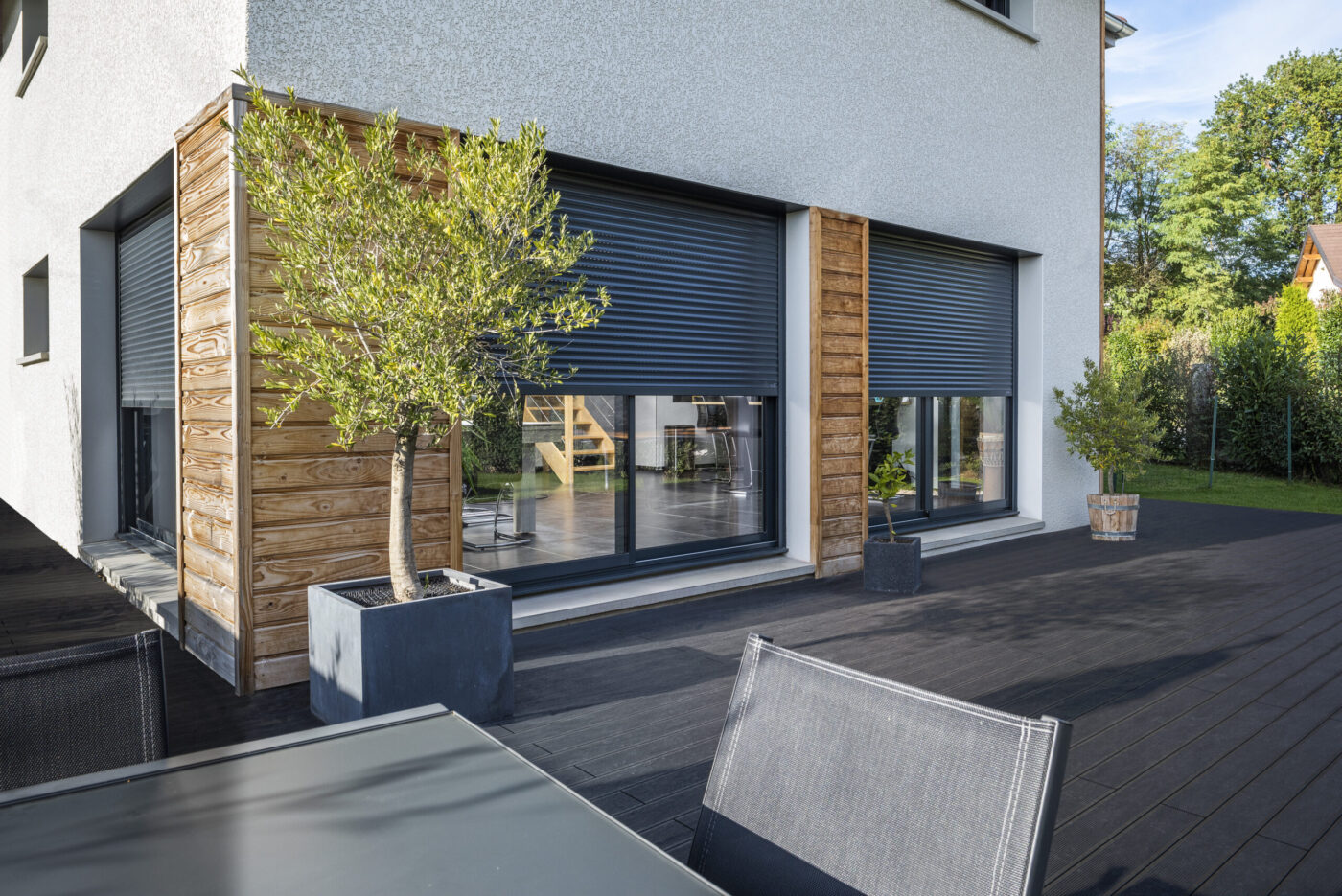 Hausfassade mit geschlossenen anthrazitfarbenen Rollläden, Holzverkleidung und Terrasse mit Olivenbaum