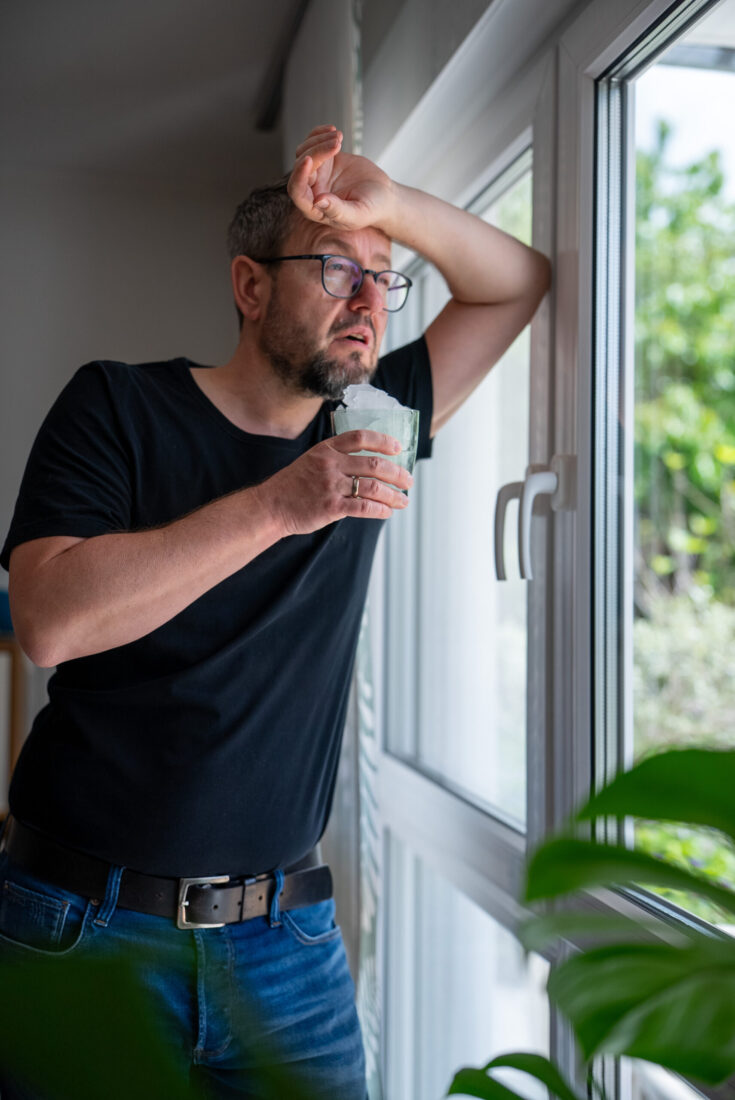 Mann hält sich erschöpft von Hitze ein Eiswasser-Glas ans Gesicht und lehnt am Fenster