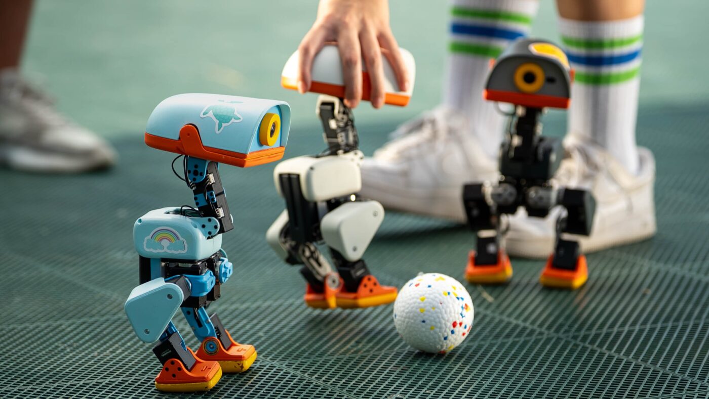 Drei Microduck-Roboter in Hellblau, Weiß und Dunkelgrau neben einem Ball auf einem grünen Sportboden