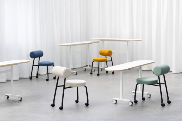 Mehrere Studiotools HighLow Chairs in Weiß, Blau, Gelb und Grau neben schmalen Tischen in einem hellen Raum